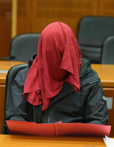 A man sits in a court room with a red cloth over his head.