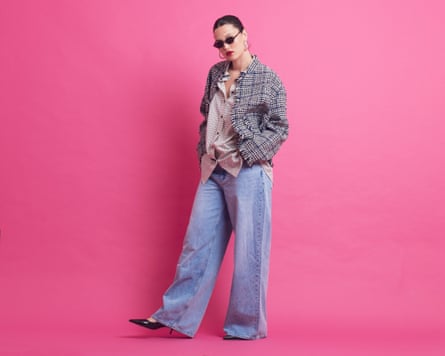 A model with dark hair in a ponytail, wearing sunglasses, red lipstick, hoop earrings, a boucle jacket, a check shirt, loose pale blue jeans and high pointed shoes.