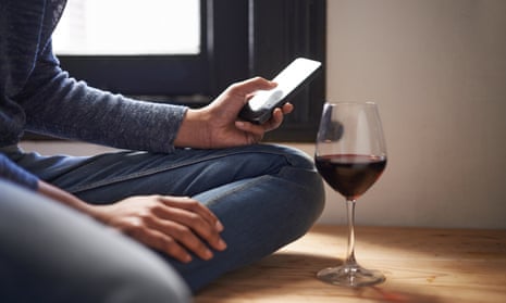 A woman’s arm and hand can be seen holding a mobile phone as she with her knee bent on a wooden bench with a glass of red wine beside her