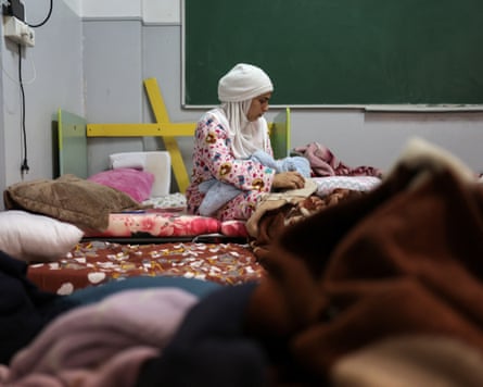 A room in a school filled with beds. A woman sits on one of the beds holding her baby