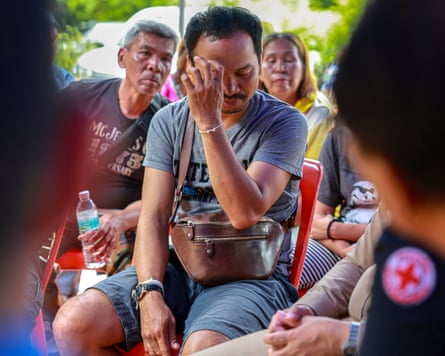 Komsan Prachan sits among a group of people; one looking on has a red cross badge on their shoulder; he holds his hand to his face and looks down.