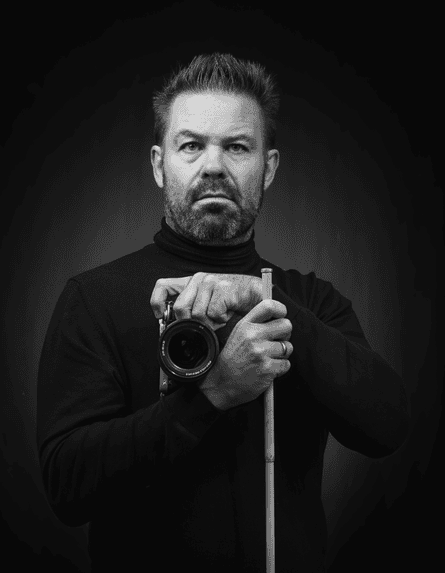 Black and white self-portrait by Gary Williamson, holding a stick in one hand and his camera in the other