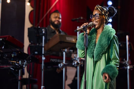 A woman in a vibrant green coat with a big fur collar and cuffs and a head scarf and wearing big spectacles sings into a microphone with a keyboard player in the background