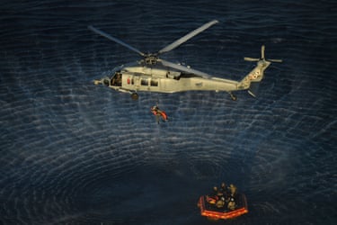 Helikopter turuncu cankurtaran botunun üzerinde havada asılı duruyor.