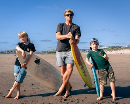 A dad and two boys lean on their surfboards on the beach. All are wearing long shorts