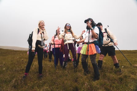 Group of women some in tutus, shorts and leggings, singing as they walk across a Scottish bog