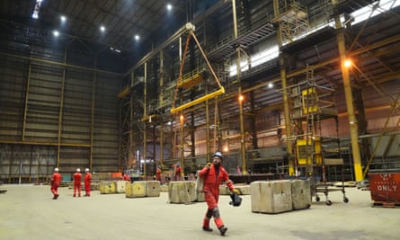 workers in orange overalls and hard hats inside a large construction hall with lifting machinery seen in the background