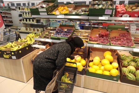 A woman leans over a crate of yellow melons in a supermarket. She has bananas in her shopping basket. There are plums, kiwis and other fruits on the shelves.