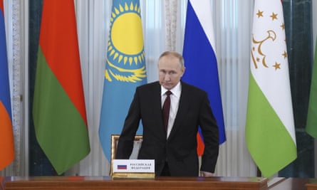 Putin is about to sit at a desk with different flags behind him