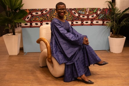 A smiling African woman in a long robe sits on a vaguely snail-shaped chair