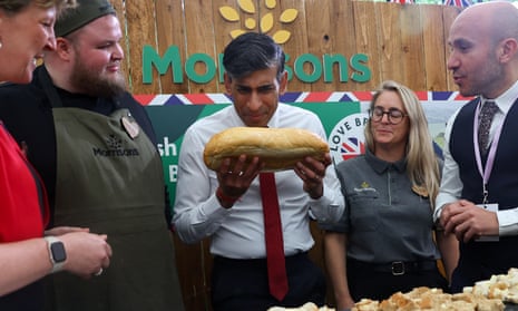 Rishi Sunak appears to be sniffing a loaf of bread he is holding in both hands, as staff from the supermarket chain Morrisons look on