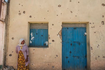 A middle-aged woman in a headscarf leans against the wall of a house pockmarked by bullets and shrapnel 
