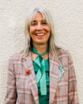 A portrait from waist up of a woman with ash blond hair and a pink checked jacket and emerald green top and neck tie standing against a wall