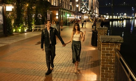 A movie still showing a white man and a white woman holding hands as they walk along a nighttime cobblestone street below streetlights.