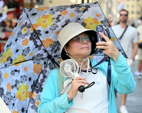 A tourist wearing a hat and holding a fan shields herself from the sun under an umbrella as she walks in Florence, Italy.