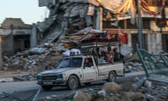 Palestinians travel in a heavily loaded car with destroyed buildings in the background and rubble in the foreground