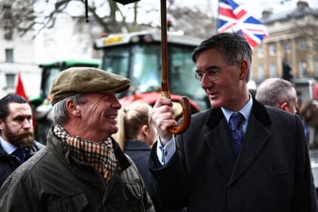 Nigel Farage (L) and Jacob Rees-Mogg chatting under an umbrella held by the latter with people and a tractor in the background
