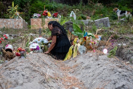 A woman in black kneels between two mounds of earth with flowers and plants at their heads.