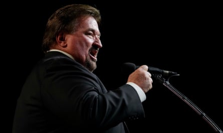 A large white man with brown hair, goatee, and black suit appears to be speaking with emphasis into a microphone on a dark stage.