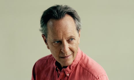 Head shot of actor Richard E Grant in red shirt against green background