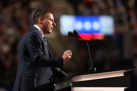 A man in a suit speaks on stage into a microphone.