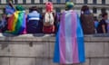 Young people wrapped in gay and trans pride flags sit on a wall with their backs to the camera