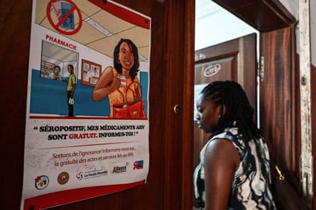 An African woman passing a poster with a drawing of a woman holding a packet of tablets at a pharmacy and a line through a picture of banknotes