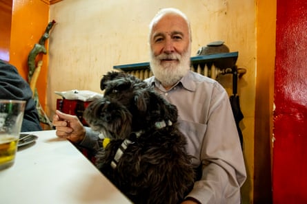 Nontas Papayiannis sits at a table with a dark grey, fluffy dog in his lap. He has white hair and a white beard. The wall behind him is painted cream, egg-yolk yellow and red.