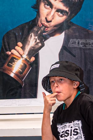 A boy wearing a bucket hat imitating Liam Gallagher’s pose on a poster behind.