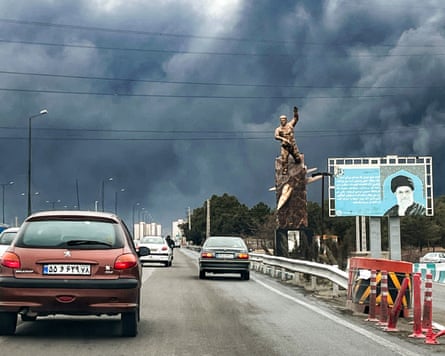 Cars on a motorway drive towards a thick smoky skyline past a war memorial statue and a billboard depicting the late Ayatollah Ali Khamenei