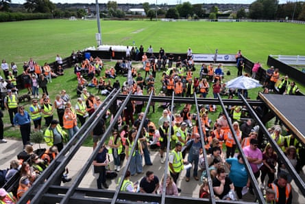 People, mostly wearing hi-vis vests, gather and listen at buildings at the edge of a recreation ground.