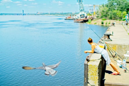 A woman leans over a concrete wall towards a large expanse of water, fishing rods propped up next to her. In the background are partially beached boats.