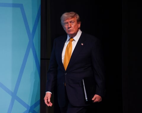 Donald Trump in black suit and yellow tie, walking onto a stage holding a leather folder.