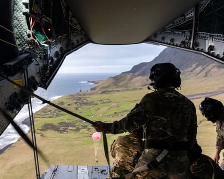 People in the open back of a military plane watch kit descending under a red parachute