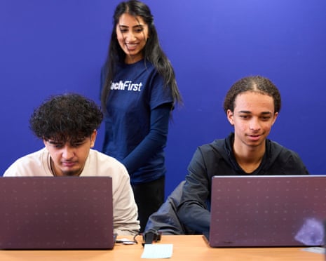 Two teenage boys sit looking at laptop screens as a young woman in a blue TechFirst T-shirt stands over them. They are all smiling. The wall behind them is deep blue.