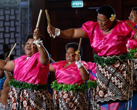 Women in pink tops and traditional Fijian costume hold up wooden sticks as they dance
