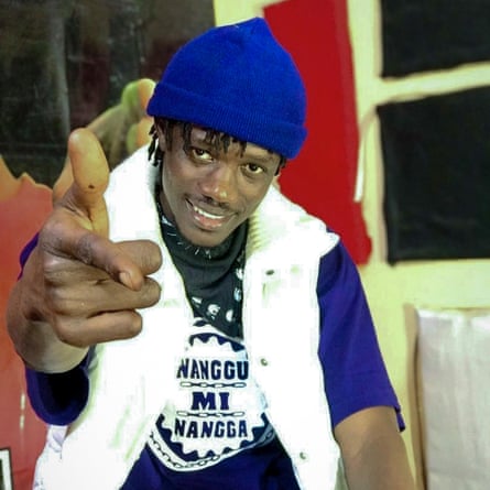 A young black man with dreadlocks seen coming out of a blue woolly hat smiles as he points at the camera