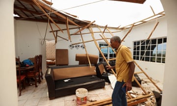 An older Black man in a yellow polo shirt and black pants walks across a tile-floored room with a couch beneath a bamboo roof and open sky.
