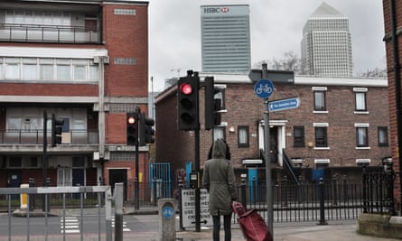 A woman photographed from behind pulling a shopping trolley at a pedestrian crossing, with two large skyscrapers behind the low-rise urban street