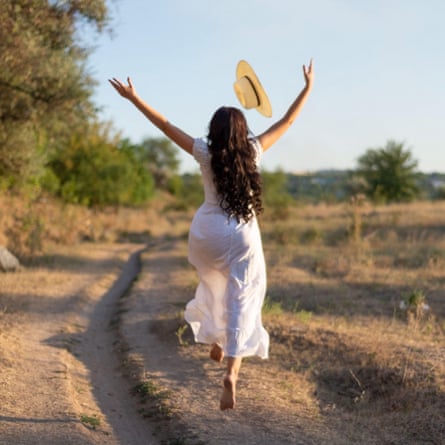 Woman leaps joyfully into the air on a country track in the sunshine, in floaty dress and straw hat