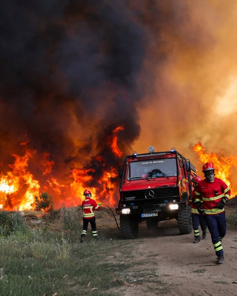 Firefighters run towards a fire truck as they prepare to tackle a wildfire in the background further down a dirt road