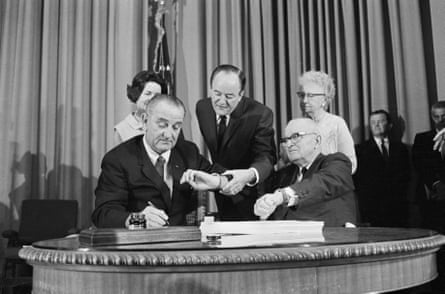 Black-and-white image of three men at a table, with two women standing by. One is signing a paper, with two looking at watches.