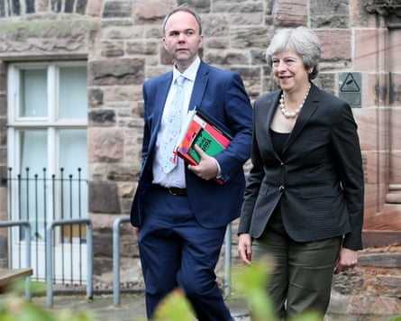 Theresa May and Gavin Barwell walk together in front of a stone building. He is carrying books and notebooks under one arm.