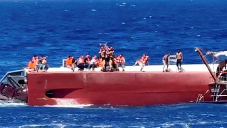 A passenger is brought aboard a rescue boat, with others wearing lifejackets sitting on deck