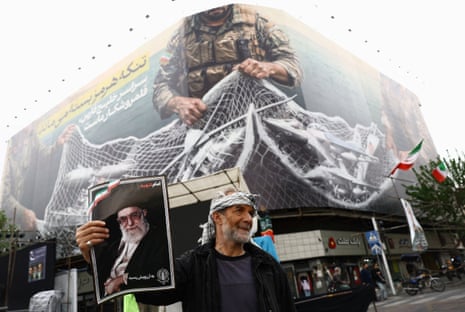 Ένας άνδρας στέκεται μπροστά από μια τοιχογραφία κρατώντας μια φωτογραφία του Αλί Χαμενεΐ.