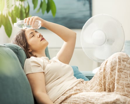 A woman sitting on a sofa in front of a fan with a bottle of water held against her head.