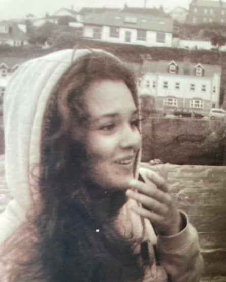 monochrome and slightly blurred picture of Maeve Boothby O’Neill smiling and holding one hand up to her face; she is outdoors with grass and houses in the background, and wears a hooded anorak