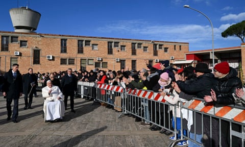 A seated pope in white robes waves at onlookers who are standing behind a barrier outside a prison. dqxikeidqkikdinv