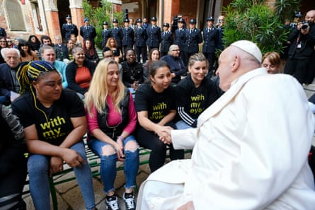 Pope Francis, dressed in white and seated in a courtyard, speaks with a group of women who sit on a bench opposite him as prison staff look on