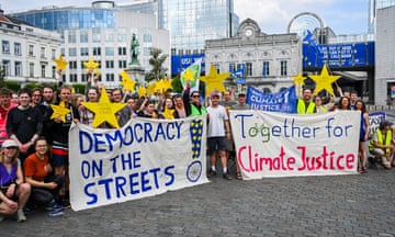 A crowd of activists stand behind two large banners that say 'democracy on the streets' and 'together for climate justice'. Some people hold large yellow stars with slogans including 'defend democracy', 'climate justice' and 'protect human rights'. Signs advertising the European elections are visible on buildings in the background